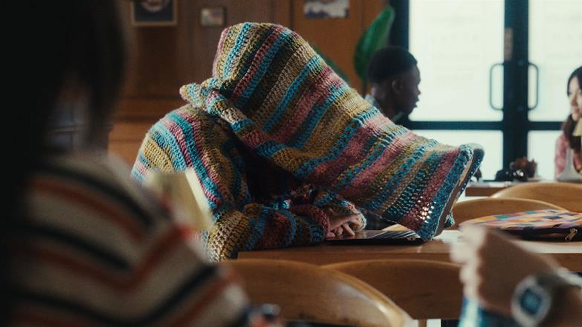 A person sits at a table wearing a colorful, oversized crocheted sweater with the hood pulled over their face. The setting appears to be a casual café.