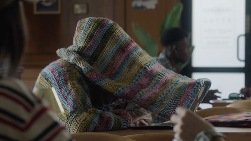 A person sits at a table wearing a colorful, oversized crocheted sweater with the hood pulled over their face. The setting appears to be a casual café.