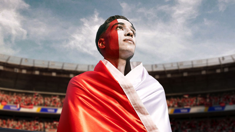 A person wrapped in a red and white flag gazes upward with a hopeful expression in a crowded stadium, under a partially cloudy sky.