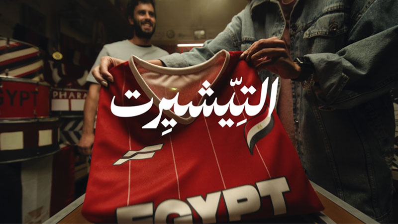Two people hold a red football jersey with "EGYPT" written on it. Bold white Arabic text overlays the image. The atmosphere appears enthusiastic and lively.