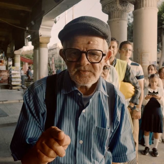 Elderly man in glasses and a flat cap pointing toward the camera, with people standing behind him outdoors