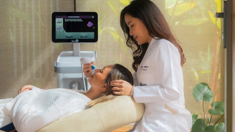 A woman in a white coat gently applies a skincare device to a reclining patient’s face in a serene clinic setting, conveying a sense of calm professionalism
