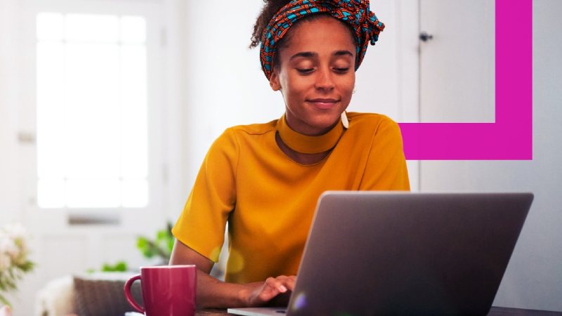 A woman in a yellow top, with a colorful headband, sits at a table working on a laptop.