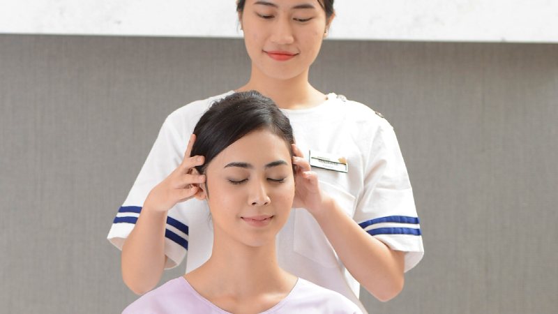 A woman in a white uniform gently massages the head of a seated woman with closed eyes, conveying relaxation and calmness in a peaceful setting.