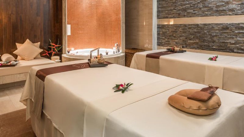 A serene spa room features two massage tables adorned with white linens and small floral arrangements, a cozy atmosphere highlighted by a warm lit Jacuzzi.