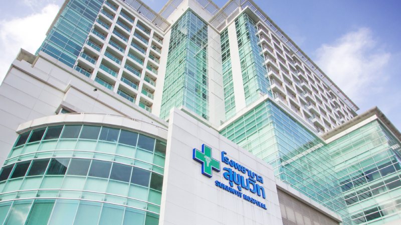 Modern hospital building with a glass facade and blue logo, viewed from a low angle against a bright sky, conveying a sense of professionalism and care