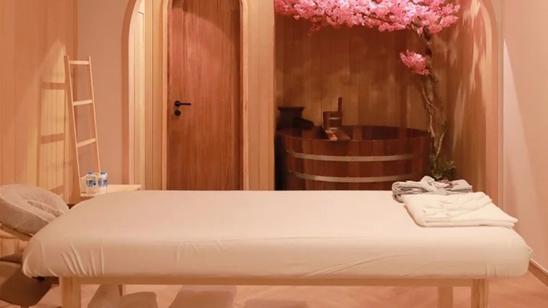 A serene spa room with a massage table, neatly folded towels, and a wooden bathtub.