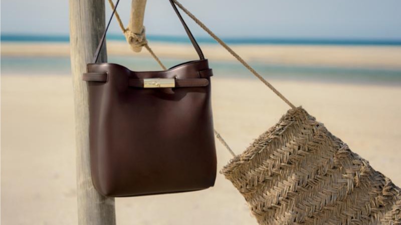 Two bags hang on a wooden post by the beach: a sleek brown leather bag and a rustic woven straw bag.