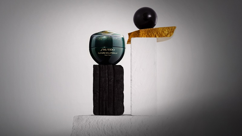 A Shiseido Future Solution LX jar is elegantly balanced on a black pedestal beside a white block with a black sphere and gold accent on top.