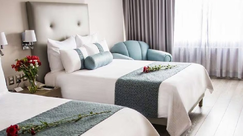 A bright hotel room with two neatly made beds, each with a rose, white and teal bedding