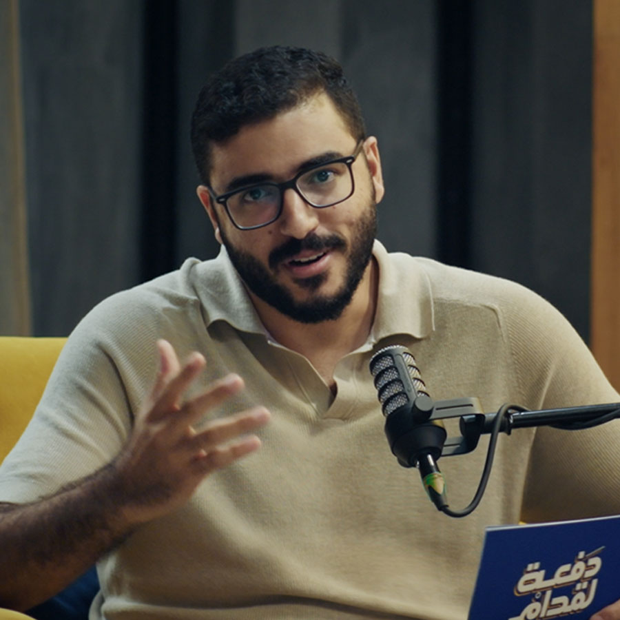 Omar Khaled seated on a yellow chair, gestures while speaking into a microphone. He holds a blue card. The setting is a cozy, modern studio.