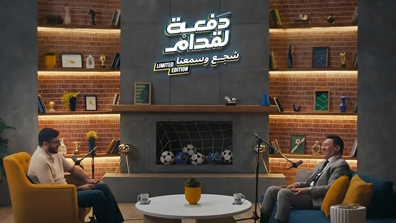 Two people are seated in a modern studio with brick walls and illuminated shelves. The shelves hold books and soccer-themed decorations. A large sign hangs overhead.