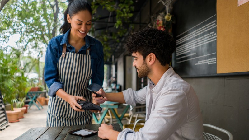A waiter holding a payment device as a man pays with his phone at an outdoor café.