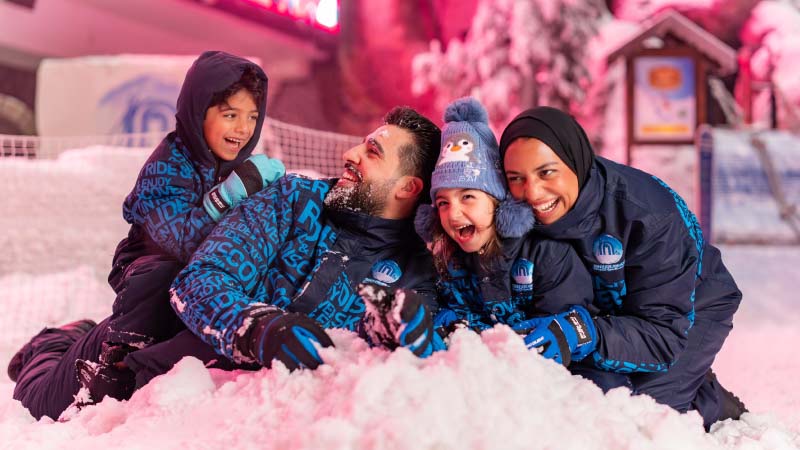 A joyful family of four in snow attire plays in the snow. They smile widely, exuding happiness. The background is warmly lit with a cozy ambiance.