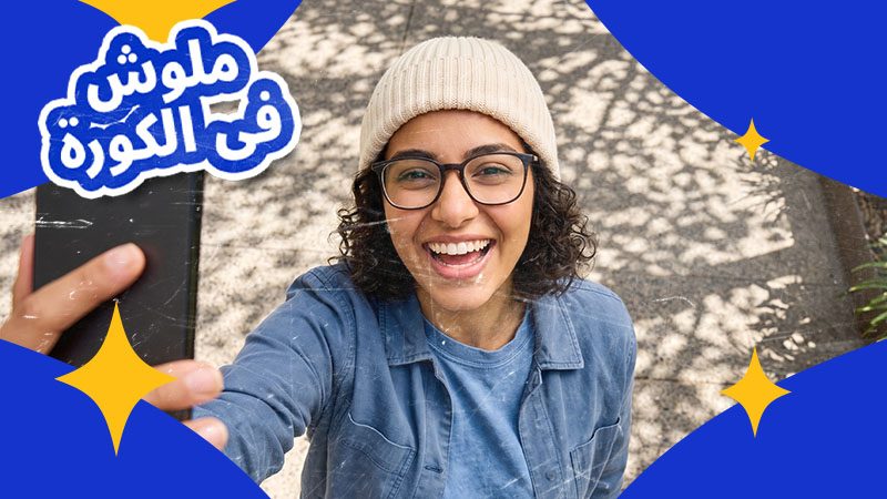 Smiling person in glasses and beanie takes a selfie. Text in Arabic is surrounded by decorative blue and yellow elements.