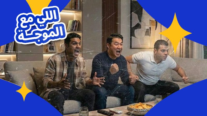 Three people on a couch excitedly watching TV, fists clenched and leaning forward. Snacks and drinks on the table. Arabic text in the upper left corner.
