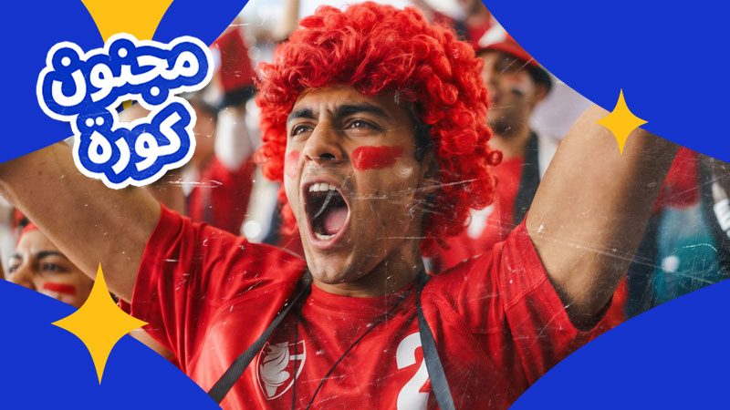 A passionate soccer fan with a red wig and face paint cheers excitedly. He wears a red jersey with "2," surrounded by a vibrant crowd, exuding enthusiasm.