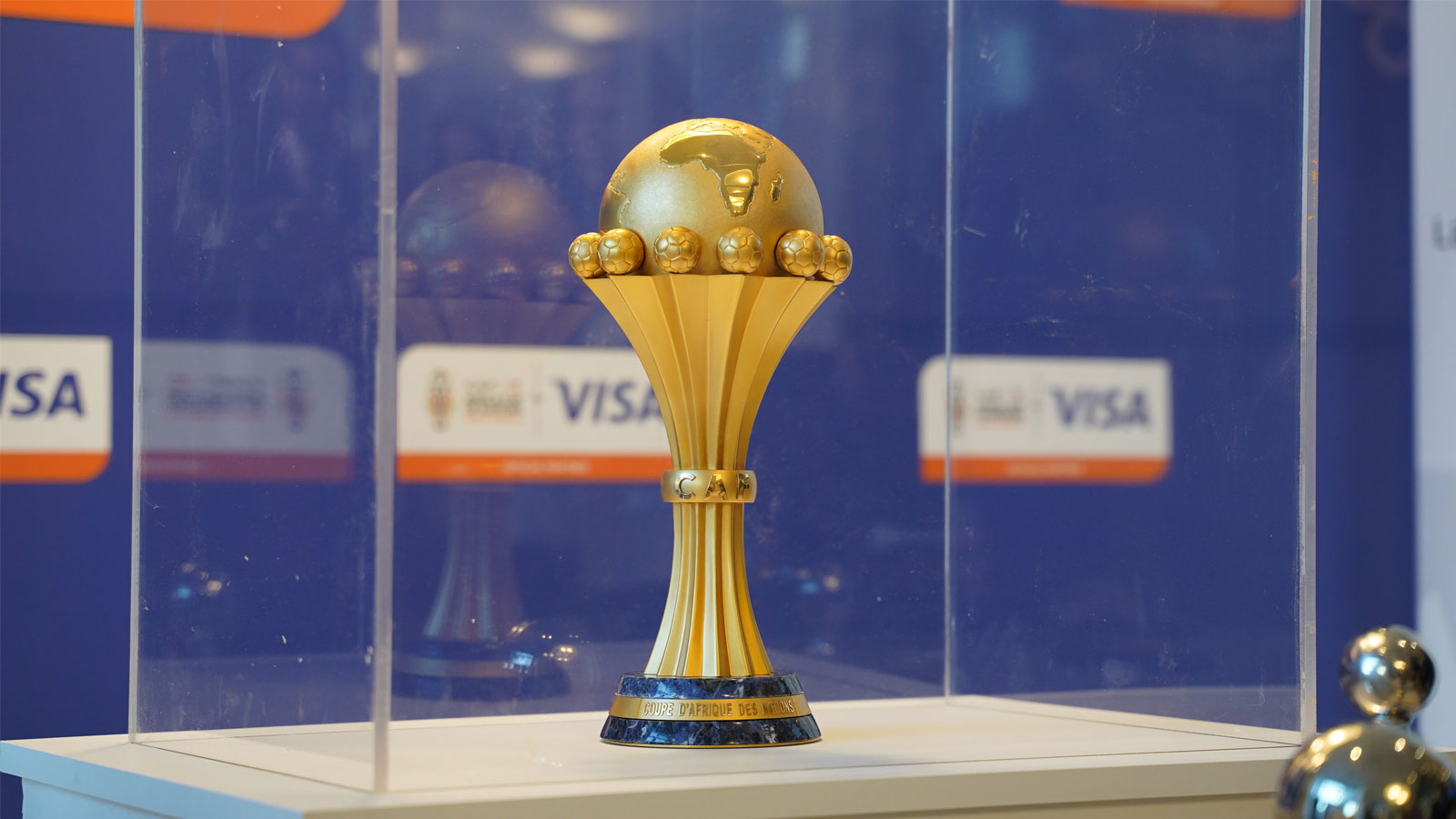 Golden trophy with globe top and six small spheres on flared base, encased in glass. Background shows sponsorship logos. Elegant and prestigious atmosphere.