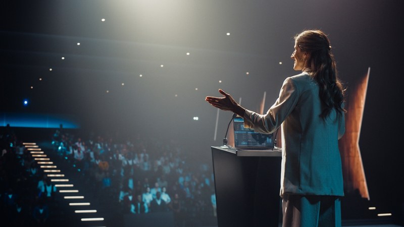A woman speaking on stage to an audience during a conference or mentorship event.