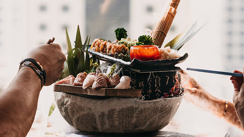 A large bowl filled with assorted sushi and sashimi, garnished with vegetables, with people using chopsticks to serve themselves.