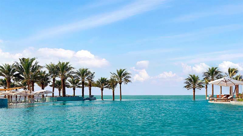 Infinity pool with clear blue water, lined with palm trees and lounge chairs, overlooking the ocean under a blue sky with scattered clouds.